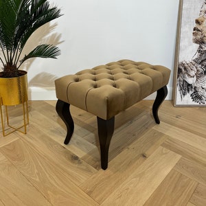 May include: A brown tufted ottoman with black wooden legs. The ottoman is sitting on a light wood floor.