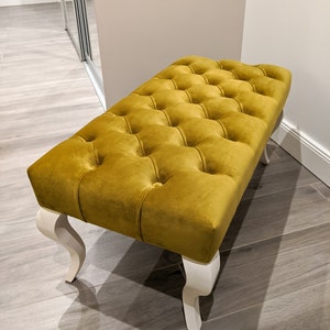 May include: A yellow velvet tufted ottoman with white legs. The ottoman has a square shape and is upholstered in a soft, plush fabric.