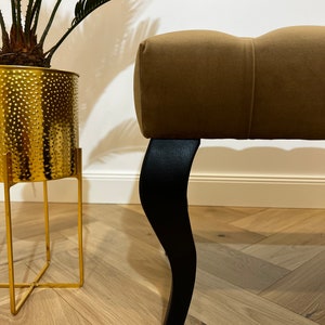 May include: A brown upholstered bench with black wooden legs. The bench has a tufted design and is sitting on a wooden floor.