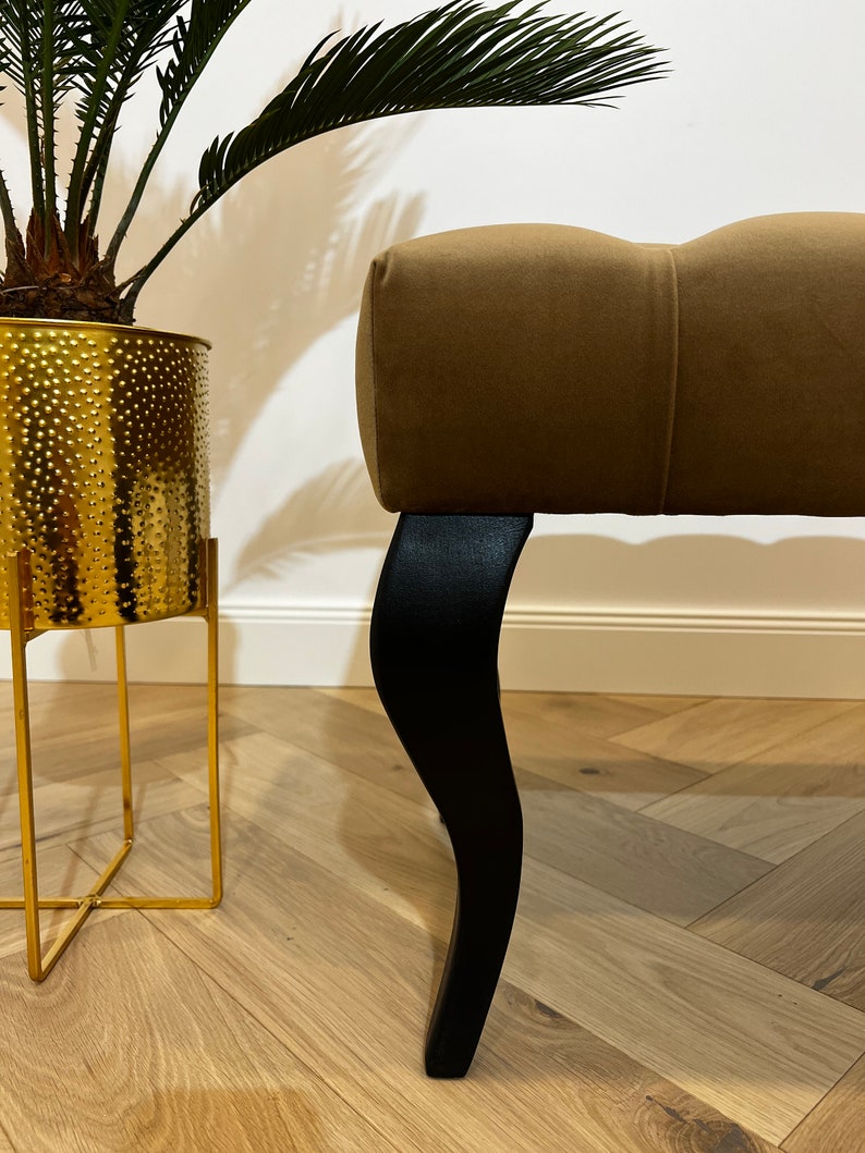 May include: A brown upholstered bench with black wooden legs. The bench has a tufted design and is sitting on a wooden floor.