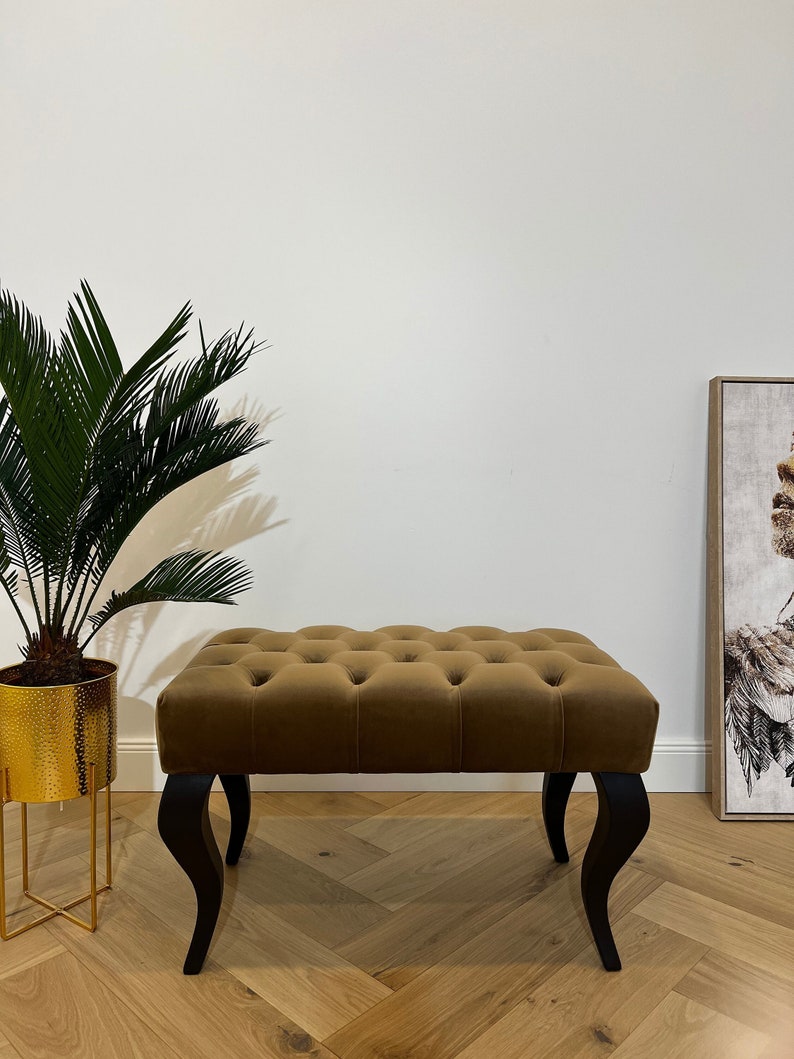 May include: A brown velvet tufted bench with black wooden legs. The bench is sitting on a wood floor with a herringbone pattern.