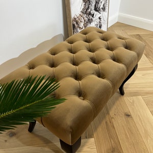 May include: A brown tufted ottoman with black wooden legs. The ottoman is upholstered in a soft velvet fabric.