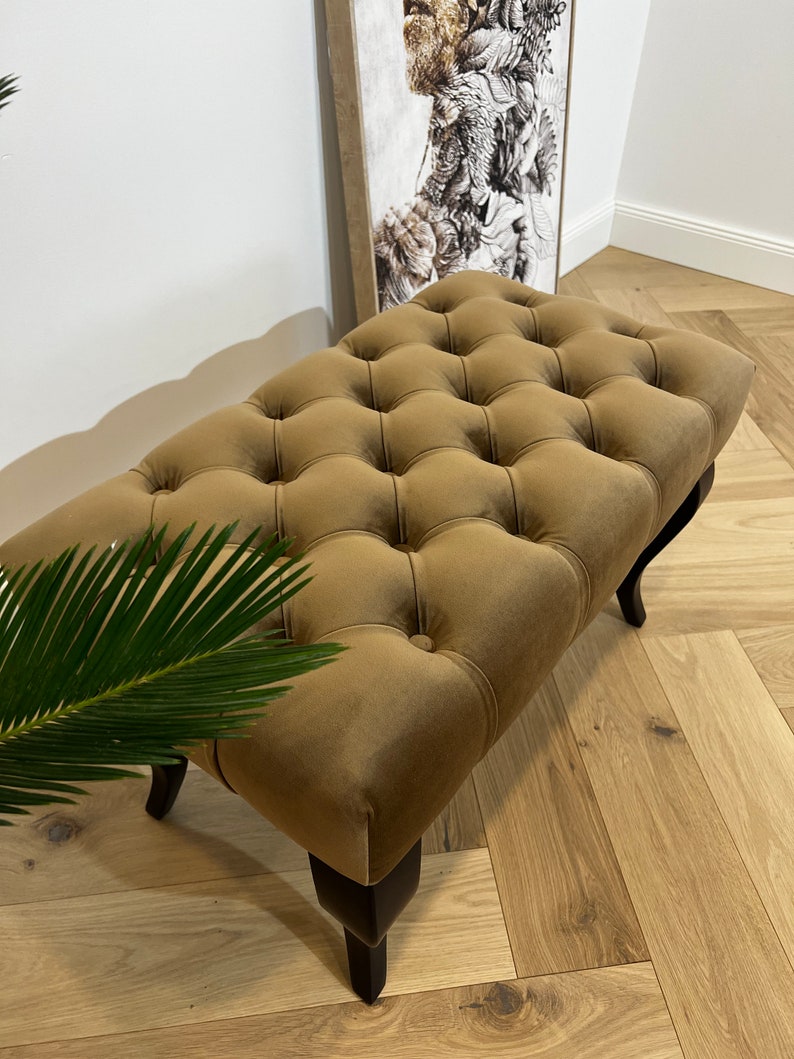 May include: A brown tufted ottoman with black wooden legs. The ottoman is upholstered in a soft velvet fabric.