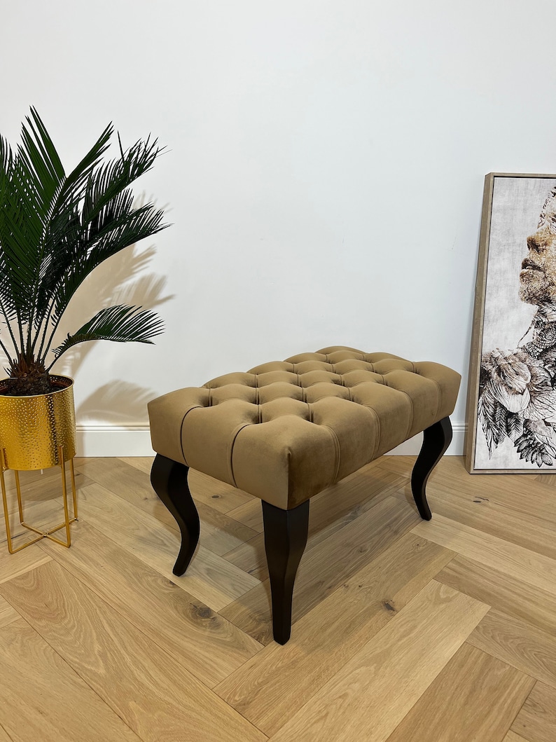 May include: A brown tufted ottoman with black wooden legs. The ottoman is sitting on a light wood floor.