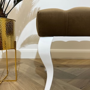 May include: A brown velvet upholstered bench with white, curved legs. The bench is sitting on a wooden floor.