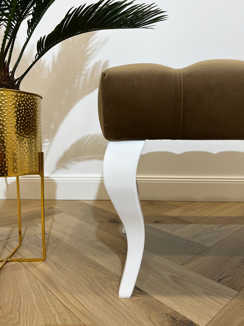 May include: A brown velvet upholstered bench with white, curved legs. The bench is sitting on a wooden floor.