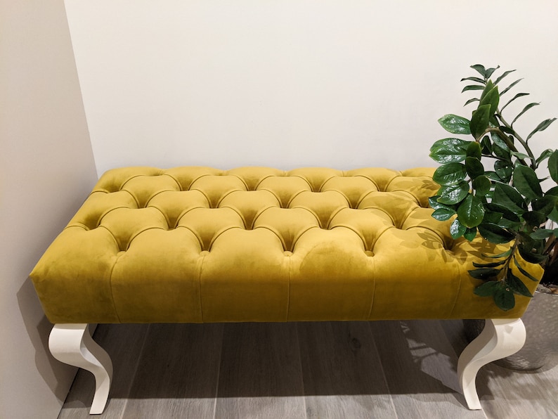May include: A yellow tufted ottoman with white legs. The ottoman has a button-tufted design and is made of a soft, plush fabric.