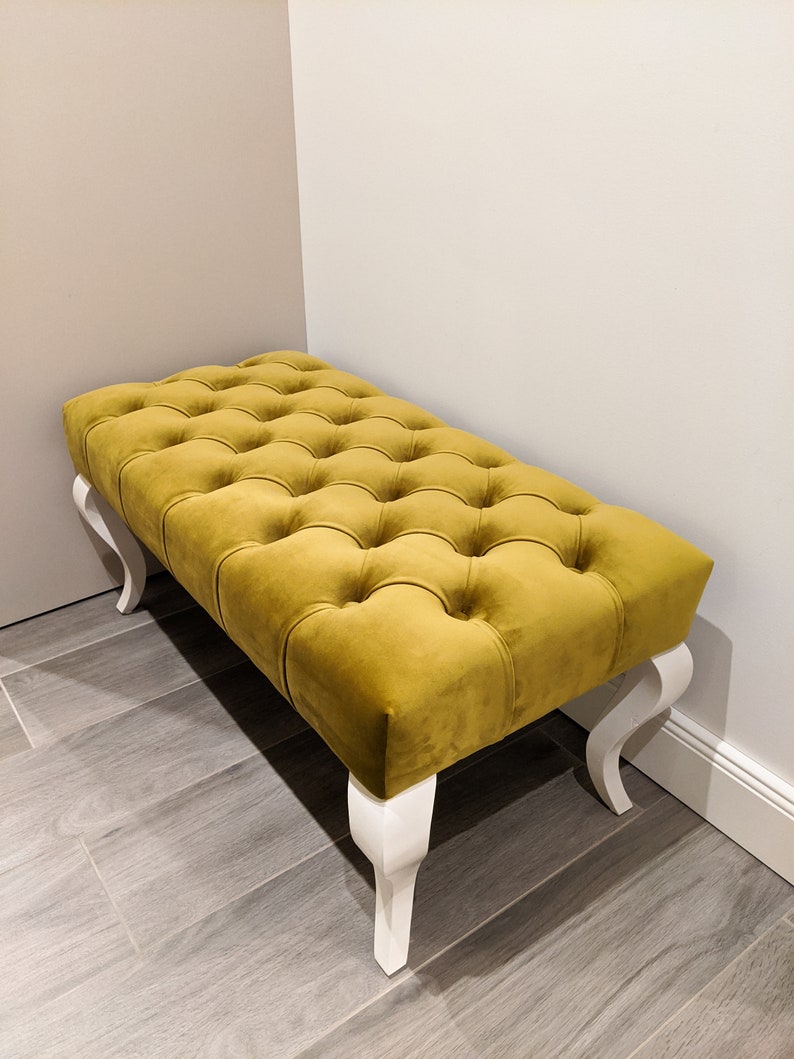 May include: A mustard yellow tufted bench with white legs. The bench is in a room with light brown wood flooring.