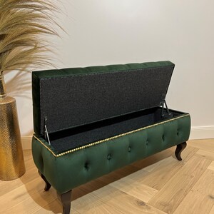 Op de afbeelding: Een groene fluwelen opbergbank met gouden bies en knoopvormige stiksels. De deksel is open, waardoor een grijze stoffen voering zichtbaar is.