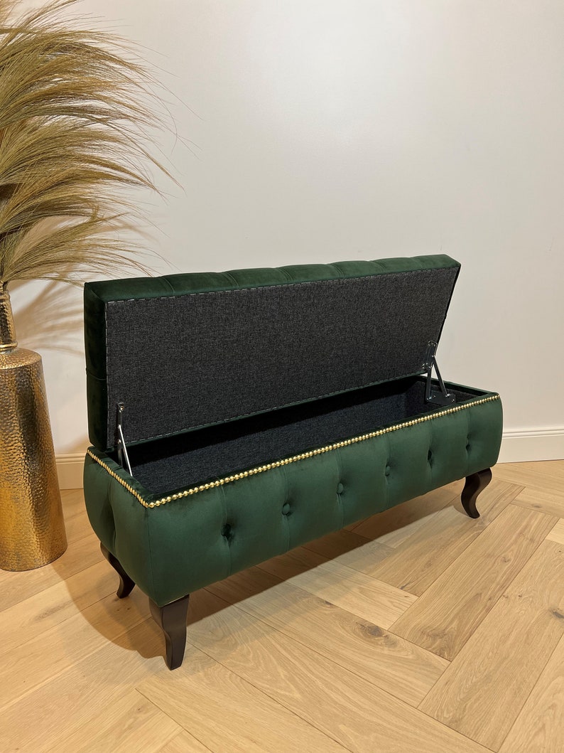 Op de afbeelding: Een groene fluwelen opbergbank met gouden bies en knoopvormige stiksels. De deksel is open, waardoor een grijze stoffen voering zichtbaar is.