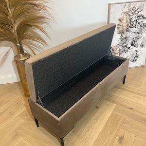 May include: A beige upholstered storage bench with a black fabric lining. The bench has a hinged lid that is open, revealing the storage space inside. The bench has black metal legs.
