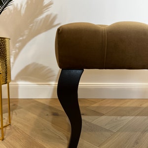 May include: A brown tufted ottoman with black wooden legs. The ottoman is sitting on a light wood floor.