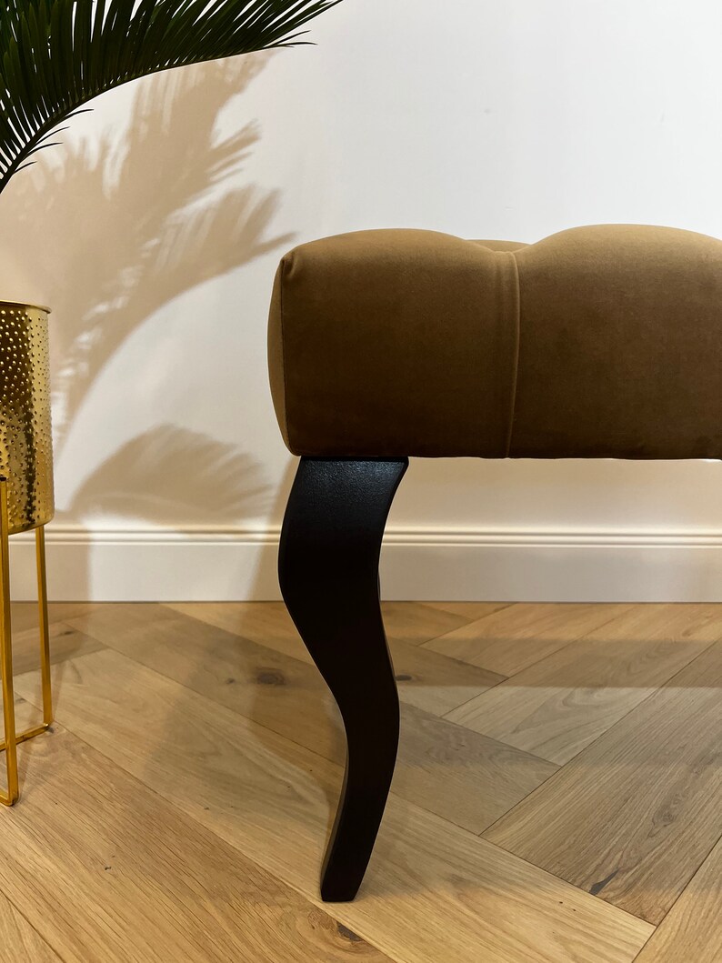 May include: A brown tufted ottoman with black wooden legs. The ottoman is sitting on a light wood floor.