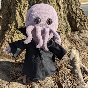 Puede incluir: Un pulpo de peluche morado que lleva un abrigo negro con ribetes dorados. El pulpo tiene grandes ojos negros y está de pie sobre una superficie marrón.