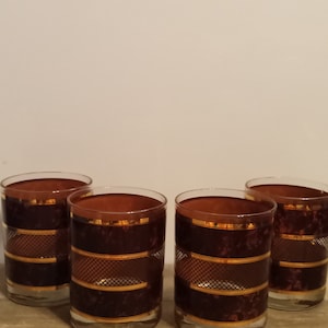 May include: Set of four brown glass tumblers with gold trim and a geometric pattern. The glasses have a clear base and a brown rim.