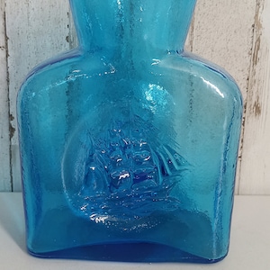 Puede incluir: Una jarra de vidrio azul turquesa con una superficie texturizada y un diseño en relieve de un barco en la parte delantera.