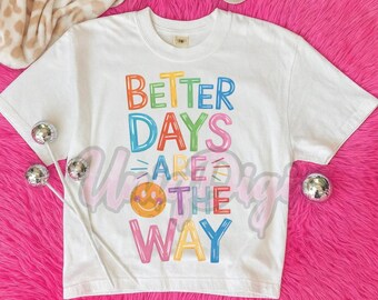 May include: White t-shirt with the text "Better Days Are The Way" in colorful block letters. The design includes a smiling face and is set against a pink fluffy background. Decorative silver ball ornaments are scattered around the shirt.