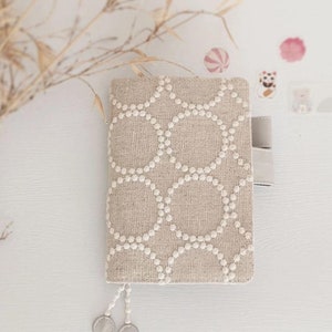 以下が含まれることがあります： 白い丸模様が施されたベージュのリネン製のノートカバー。カバーにはシルバーのジッパーとシルバーのタッセルが付いています。カバーには「Open to customisation」という文字が印刷されています。カバーの下には「A5 / A6 / B6 / 5-Year / Weeks / TN」という文字が印刷されています。