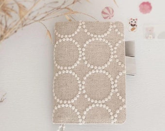 May include: A beige linen notebook cover with a white circle pattern. The cover has a silver zipper pull and a silver tassel. The text "Open to customisation" is printed on the cover. The text "A5 / A6 / B6 / 5-Year / Weeks / TN" is printed below the cover.