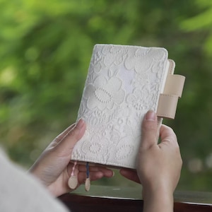 May include: Cream-colored journal with a floral lace pattern. The journal has a leather strap closure and decorative tassels. The cover is a soft, off-white color. The journal is held in two hands against a blurred green background.