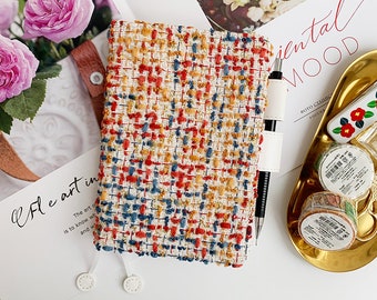 Op de afbeelding: Een veelkleurige geweven notitieboekhoes met een pen vastgezet met een witte leren riem. De hoes heeft rode, blauwe en gele draden. Het notitieboek ligt naast een gouden dienblad met decoratieve items en een bloemstuk.