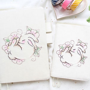 May include: Two off-white journals with a bunny and floral embroidery design. The larger journal has a tab closure. The bunny is white with pink accents, surrounded by pink flowers and green leaves. The smaller journal has a similar design.