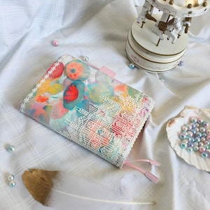 May include: A pink and white fabric planner cover with a watercolour design of flowers and a city skyline. The cover has a white lace trim and pink ribbon ties.
