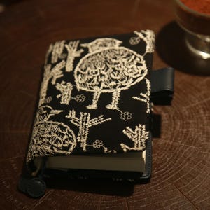 May include: Black journal cover with white embroidered tree and floral designs. The cover has a navy blue leather strap and a matching closure. The journal is closed and resting on a wooden surface.