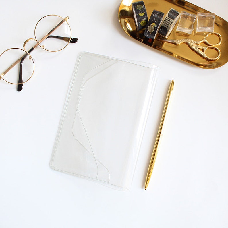 Puede incluir: Una cubierta de cuaderno de pl&aacute;stico transparente con un bol&iacute;grafo dorado sobre una superficie blanca.