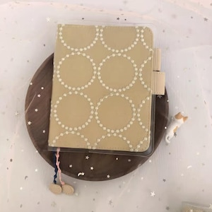Puede incluir: Una cubierta de cuaderno de tela beige con un patr&oacute;n de c&iacute;rculos blancos. La cubierta est&aacute; en una funda de pl&aacute;stico transparente. El cuaderno tiene un borla azul y rosa.