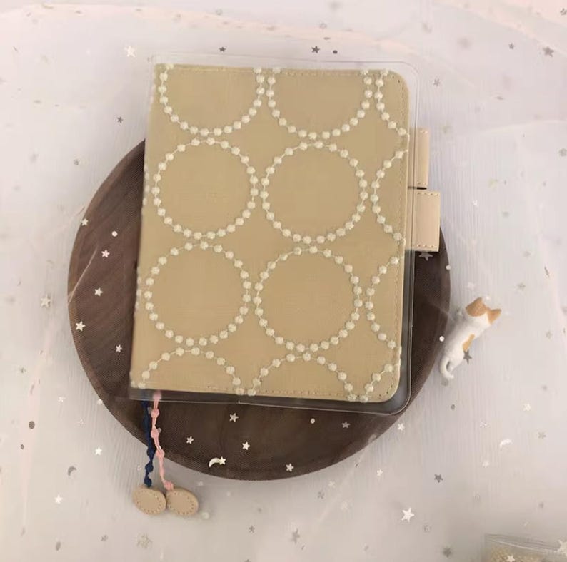 Puede incluir: Una cubierta de cuaderno de tela beige con un patr&oacute;n de c&iacute;rculos blancos. La cubierta est&aacute; en una funda de pl&aacute;stico transparente. El cuaderno tiene un borla azul y rosa.