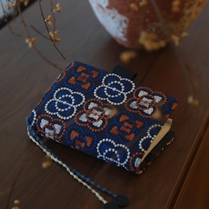 May include: A navy blue book cover with an embroidered floral pattern in white and orange. The cover has a black strap closure and a cream-colored tassel with black beads. The book cover is on a wooden surface.