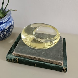May include: A pale yellow, circular glass bowl with a smooth, rounded exterior and a recessed center. The glass is translucent, allowing light to pass through its thick, curved form, creating soft reflections and a subtle, warm, golden hue.