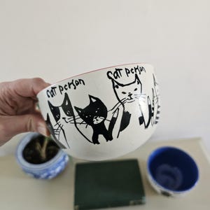 Puede incluir: Un cuenco de cer&aacute;mica blanco con borde rojo, con ilustraciones de gatos negros y el texto "Cat person". El cuenco est&aacute; sostenido por una mano, con una peque&ntilde;a maceta azul y blanca y un peque&ntilde;o cuenco azul en el fondo.