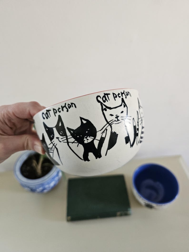 Puede incluir: Un cuenco de cer&aacute;mica blanco con borde rojo, con ilustraciones de gatos negros y el texto "Cat person". El cuenco est&aacute; sostenido por una mano, con una peque&ntilde;a maceta azul y blanca y un peque&ntilde;o cuenco azul en el fondo.