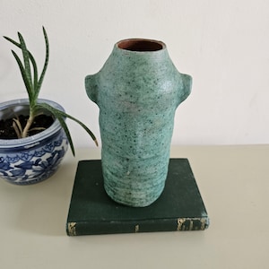 May include: A handcrafted, turquoise ceramic vase with two small handles. The vase has a textured surface and a brown interior. It is displayed on a dark green book, with a blue and white patterned pot with a plant in the background.