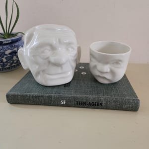 May include: Two white ceramic vessels shaped like human heads with distinct facial expressions. The larger vessel depicts an older face with deep wrinkles, while the smaller cup features a younger, frowning face. Both are resting on a gray book.