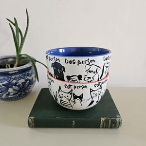 Puede incluir: Dos cuencos de cer&aacute;mica apilados con ilustraciones en blanco y negro de perros y gatos. El cuenco superior tiene un interior azul y las palabras "Dog person" escritas alrededor del borde. El cuenco inferior tiene las palabras "Cat person".