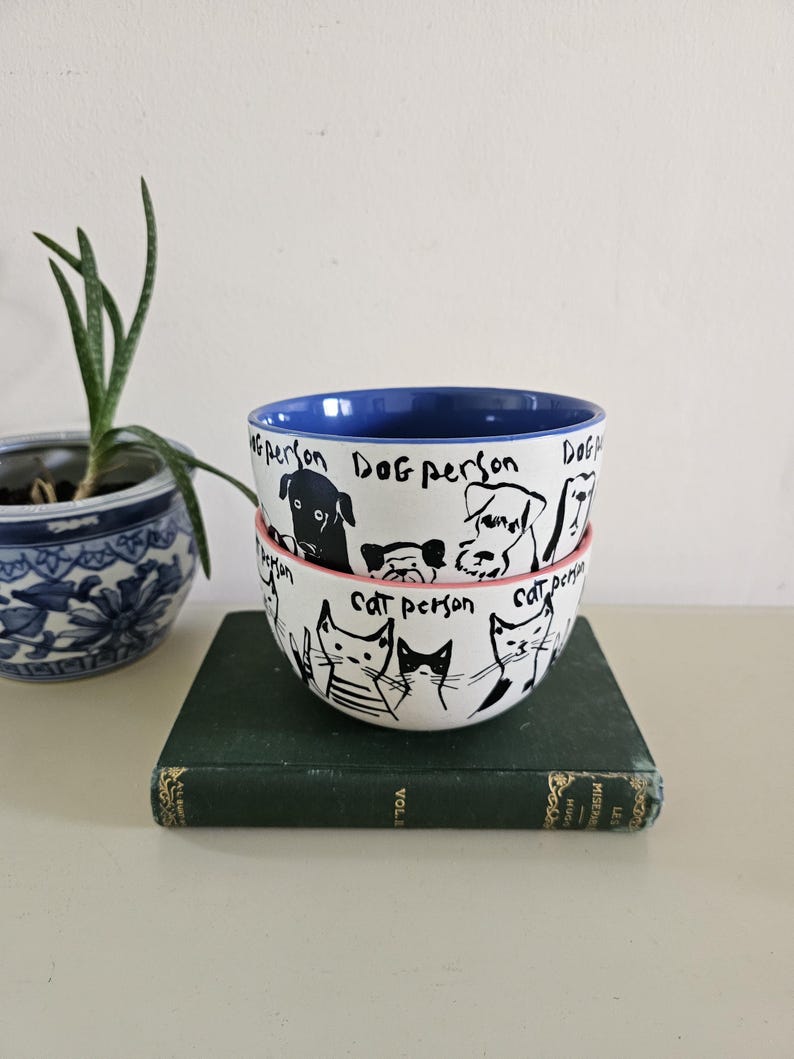 Puede incluir: Dos cuencos de cer&aacute;mica apilados con ilustraciones en blanco y negro de perros y gatos. El cuenco superior tiene un interior azul y las palabras "Dog person" escritas alrededor del borde. El cuenco inferior tiene las palabras "Cat person".