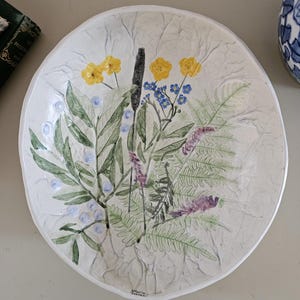 May include: A white ceramic decorative plate with a hand-painted floral design. The plate features yellow, blue, and purple flowers, green leaves, and a textured surface. The plate is round and has a slightly raised edge.