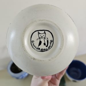 Puede incluir: Jarr&oacute;n de cer&aacute;mica color crema con forma redondeada. La base presenta una ilustraci&oacute;n de gato en blanco y negro dentro de un marco circular con la palabra "ANTHROPOLOGIE". El jarr&oacute;n es sostenido por una mano, con otros objetos de cer&aacute;mica en el fondo.