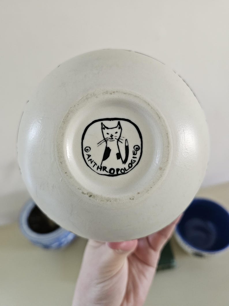 Puede incluir: Jarr&oacute;n de cer&aacute;mica color crema con forma redondeada. La base presenta una ilustraci&oacute;n de gato en blanco y negro dentro de un marco circular con la palabra "ANTHROPOLOGIE". El jarr&oacute;n es sostenido por una mano, con otros objetos de cer&aacute;mica en el fondo.