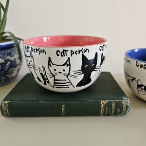 Puede incluir: Cuenco de cer&aacute;mica blanco con interior rosa, con ilustraciones de gatos negros y el texto "cat person". El cuenco est&aacute; sobre un libro verde. Una maceta con estampado azul y blanco es visible en el fondo.