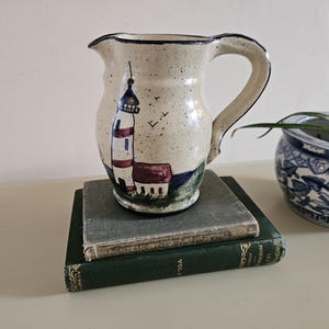 May include: A ceramic pitcher with a lighthouse design. The pitcher is off-white with a blue rim and handle. The lighthouse is painted in red and white stripes with a blue top. The pitcher is sitting on top of two books.