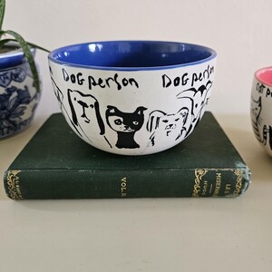 Puede incluir: Cuenco de cer&aacute;mica blanco con borde interior azul, con dibujos lineales negros de varias razas de perros. Las palabras "Dog person" est&aacute;n escritas en negro. El cuenco est&aacute; sobre un libro verde.
