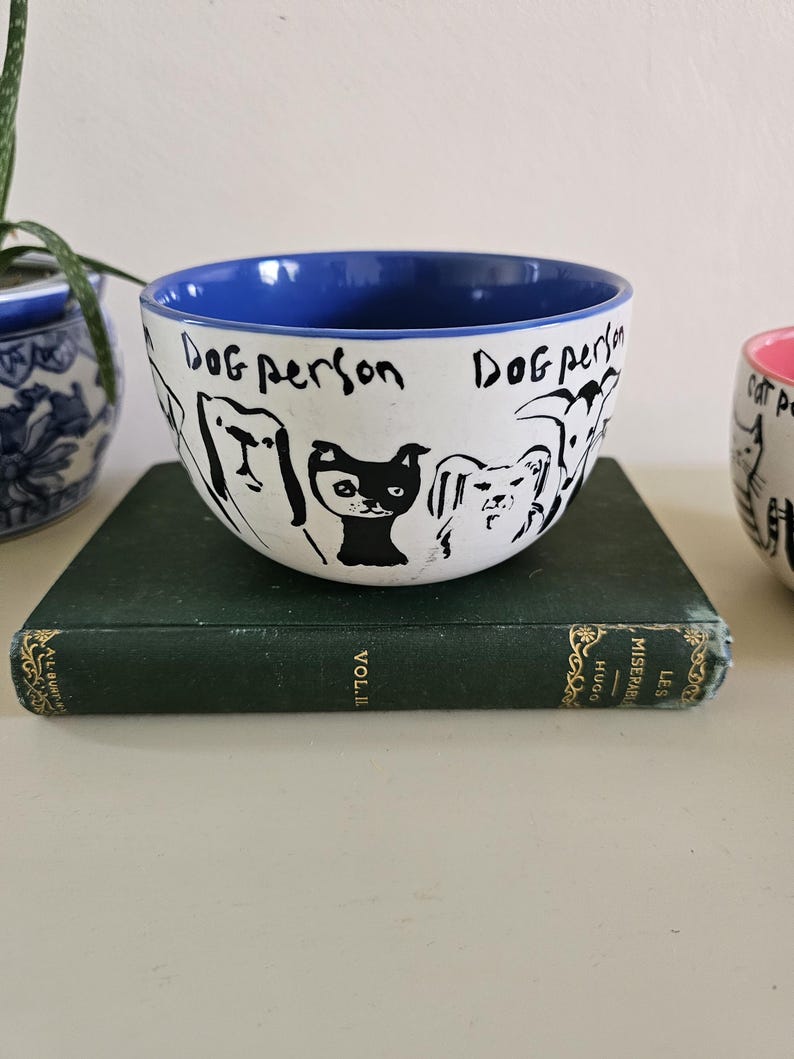 Puede incluir: Cuenco de cer&aacute;mica blanco con borde interior azul, con dibujos lineales negros de varias razas de perros. Las palabras "Dog person" est&aacute;n escritas en negro. El cuenco est&aacute; sobre un libro verde.