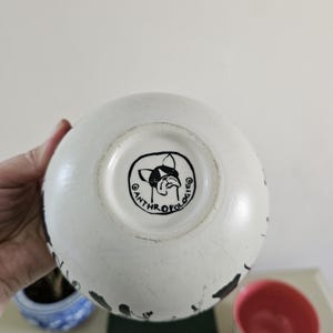 Puede incluir: Cuenco de cer&aacute;mica blanca con salpicaduras de pintura negra. El cuenco tiene un interior rosa y una ilustraci&oacute;n en blanco y negro de un perro en la base. La palabra "ANTHROPOLOGIE" est&aacute; impresa alrededor de la ilustraci&oacute;n del perro.