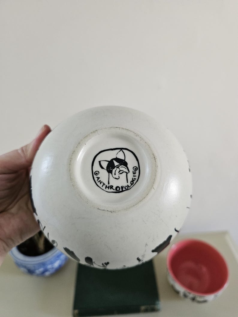 Puede incluir: Cuenco de cer&aacute;mica blanca con salpicaduras de pintura negra. El cuenco tiene un interior rosa y una ilustraci&oacute;n en blanco y negro de un perro en la base. La palabra "ANTHROPOLOGIE" est&aacute; impresa alrededor de la ilustraci&oacute;n del perro.