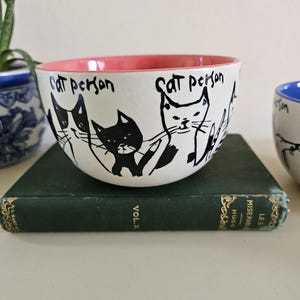 Puede incluir: Cuenco de cer&aacute;mica blanco con borde interior rosa, con ilustraciones de gatos negros y las palabras "Cat Person" escritas a mano. El cuenco est&aacute; sobre un libro verde.