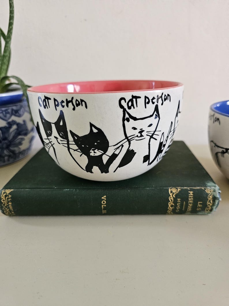 Puede incluir: Cuenco de cer&aacute;mica blanco con borde interior rosa, con ilustraciones de gatos negros y las palabras "Cat Person" escritas a mano. El cuenco est&aacute; sobre un libro verde.